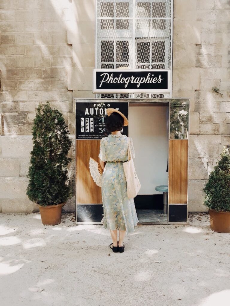 woman wearing a hat and a fan in front of an old photo booth in Arles