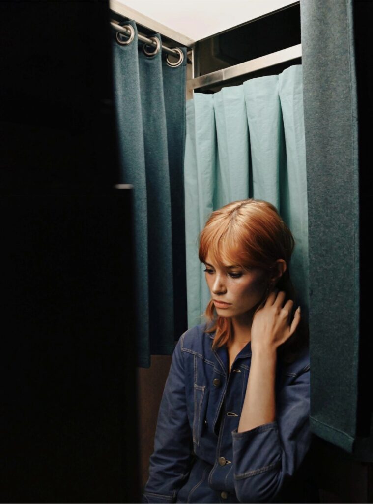 femme rousse assise dans un photomaton avec des rideaux bleu